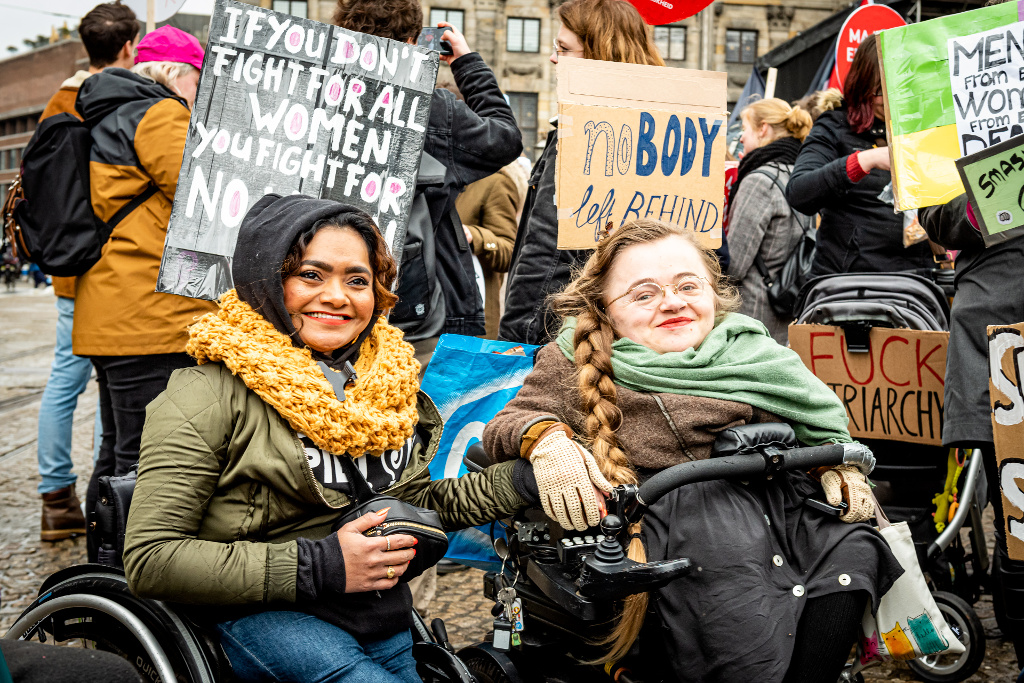 Image Description: 

A photo taken during the Women’s march 2020. On the left side of the picture sits Jeanette Chedda, who is holding the hand of Mira Thompson. Mira is sitting on the right side of the picture. Chedda is a brown woman with curly brown hair and a wide smile. She’s wearing a yellow scarf and a black hoodie. Mira is a white woman, she’s smiling at the viewer. She’s wearing her long blonde hair in a braid, the braid hangs over her green scarf. Both women are in a wheelchair. Behind them, you can see a lot of protestors and various signs they carry with them. On the sign behind Chedda is written ‘if you don’t fight for all women, you fight for no women’ and on the sign behind Mira is written ‘noBODY left behind’ with emphasis on body.
