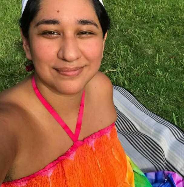 @annielainey. Annie is een bruine vrouw en zit op een deken op het gras. Ze draagt een regenboogjurk.