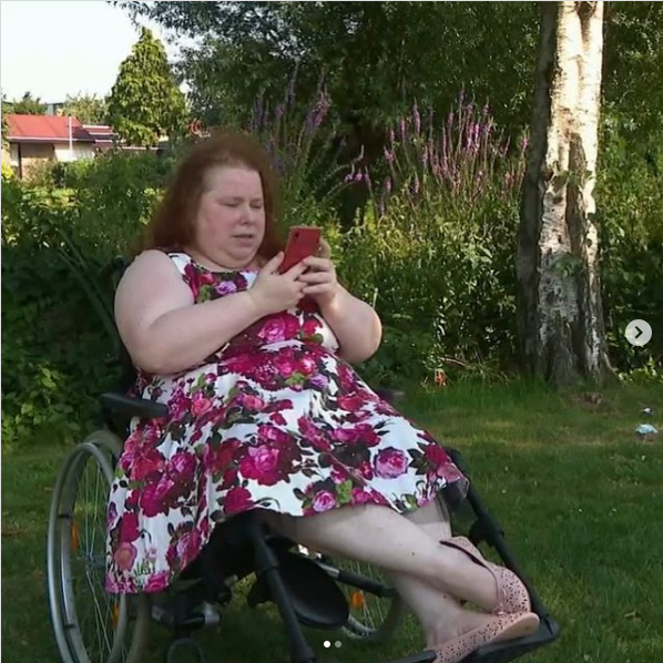 Jacquie zit in een rolstoel in een park. Hen heeft een witte jurk met roze bloemen aan en kijkt op diens telefoon.⁣