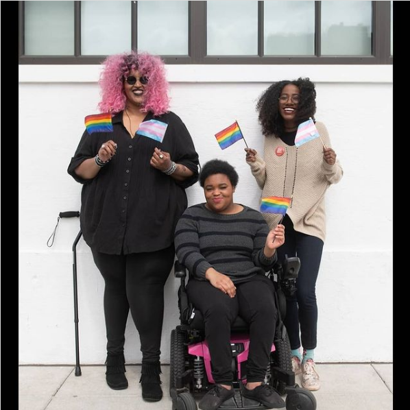 drie Zwarte gehandicapte mensen lachen en hebben kleine vlaggetjes in hun hand. Links staat een non-binair persoon, hun stok rust tegen de witte muur. hen heeft een regenboogvlag en een transgendervlag vast. In het midden zit een non-binair persoon in een rolstoel en heeft een regenboogvlag vast. rechts staat een vrouw met een regenboogvlag en een transgendervlag.
