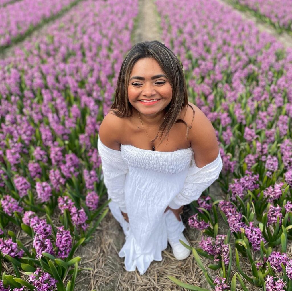 Chedda staat in een veld met paarse hyacinten. Ze draagt een witte strapless jurk en een wit vest dat haar schouders onbedekt laat. Ze lacht in de camera. Ze is een bruine vrouw met stijl donker haar. 